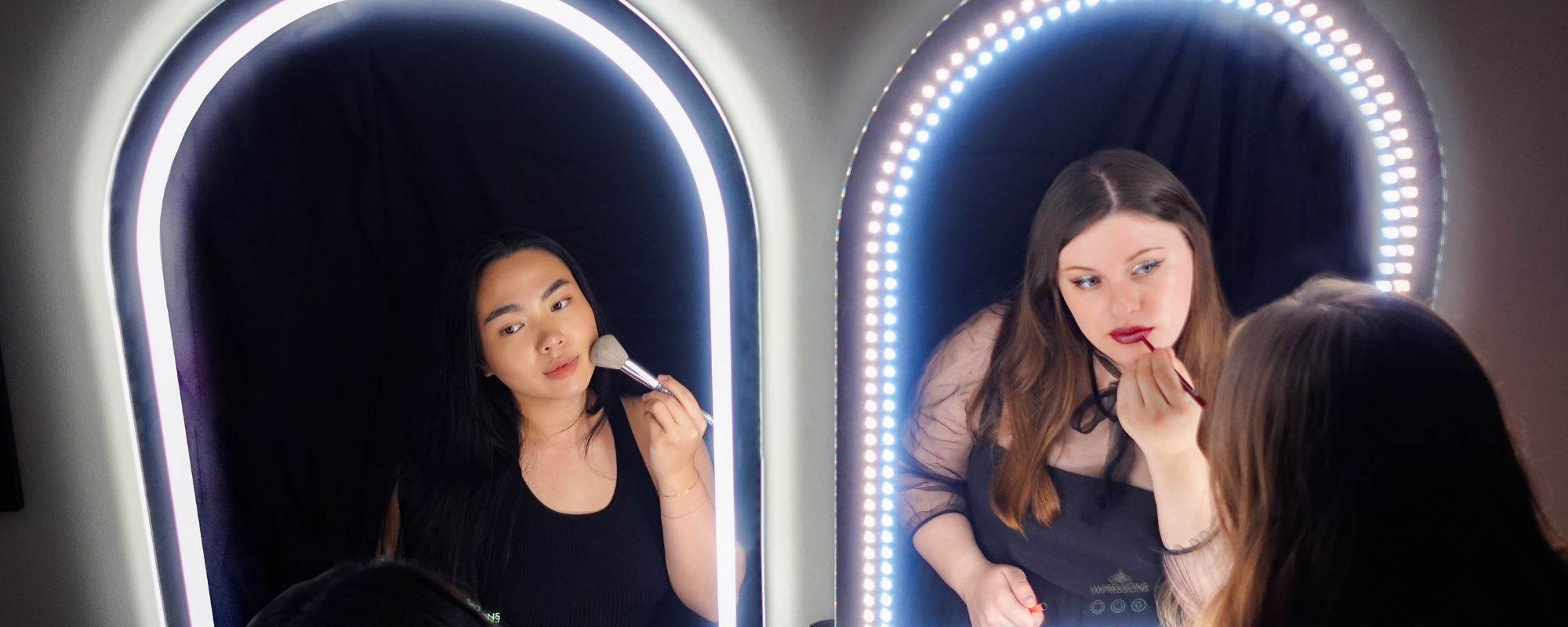 Two people applying makeup while looking into illuminated mirrors.